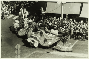 Ein Schwarz-Weiß-Foto eines mit Blumen geschmückten Umzugswagens, mit einer Gruppe von Menschen auf der Straße und einem Mast im Hintergrund.