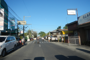 Eine belebte Stadtstraße mit Fahrzeugen, Fußgängern, Radfahrern, Gebäuden, Strommasten und Bäumen unter einem klaren blauen Himmel.