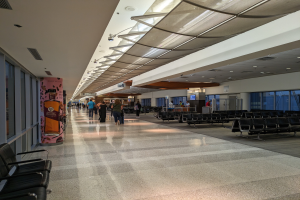 Ein langer Flughafenflur mit Menschen, die hindurchgehen, Stühle auf der rechten Seite, Glasfenster auf der linken Seite und Deckenlampen, mit ein paar Menschen im Hintergrund.