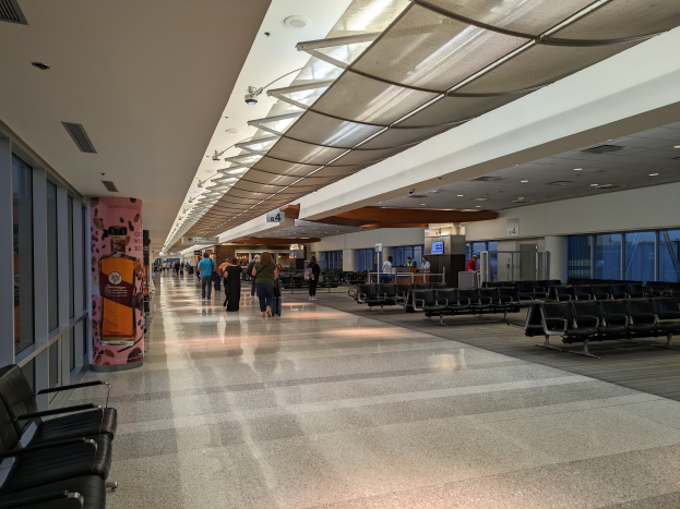 Ein langer Flughafenflur mit Menschen, die hindurchgehen, Stühle auf der rechten Seite, Glasfenster auf der linken Seite und Deckenlampen, mit ein paar Menschen im Hintergrund.