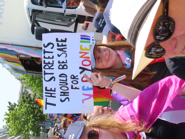 Eine Gruppe von Menschen steht vor einem Lkw und hält ein Schild hoch, auf dem "Die Straßen sollten sicher sein" steht. Einige der Menschen tragen Mützen und Schutzbrillen, andere halten Schilder. Im Hintergrund sind Bäume, Gebäude und ein klarer blauer Himmel zu sehen.