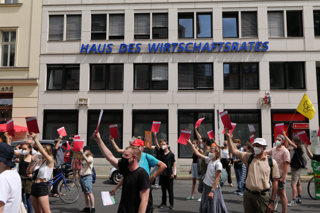Eine Gruppe von Menschen, die vor einem Gebäude protestieren und Fahnen und Schilder halten, mit Fahrzeugen auf der Straße und einer Tafel mit Text auf der rechten Seite.