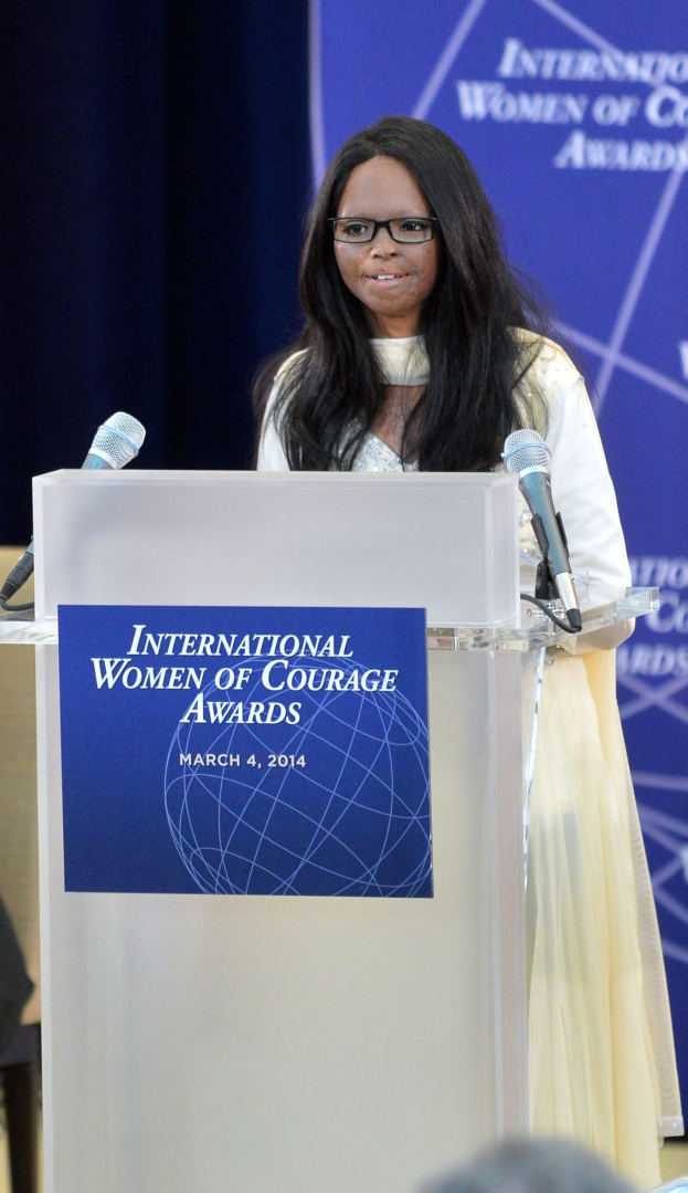 Eine Frau in einem weißen Kleid und Brille steht an einem Podium mit zwei Mikrofonen und einer Tafel und spricht vor einer Menge bei der 'International Women of Courage Awards 2014'-Veranstaltung.
