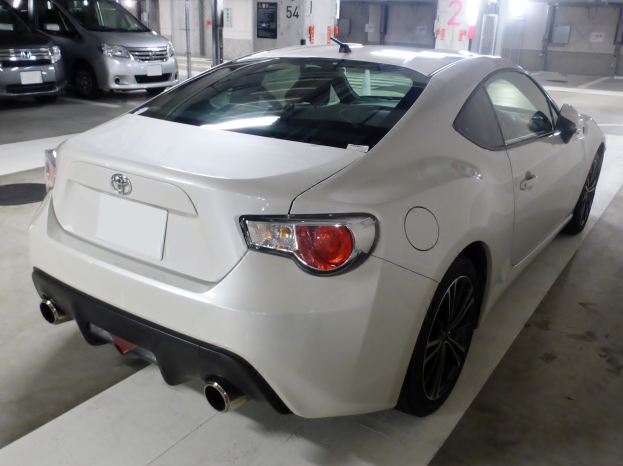 Ein weißer Sportwagen, der in einer hell erleuchteten Parkgarage neben anderen Fahrzeugen geparkt ist, mit einer Wand im Hintergrund.