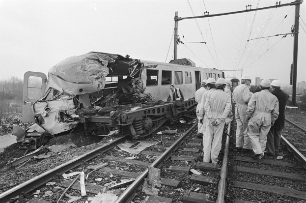 Schwarzes und weißes Bild einer Zugkollision auf den Schienen mit Menschen in Helmen in der Nähe, Strommasten, Bäume, Gebäude und einen klaren Himmel im Hintergrund.